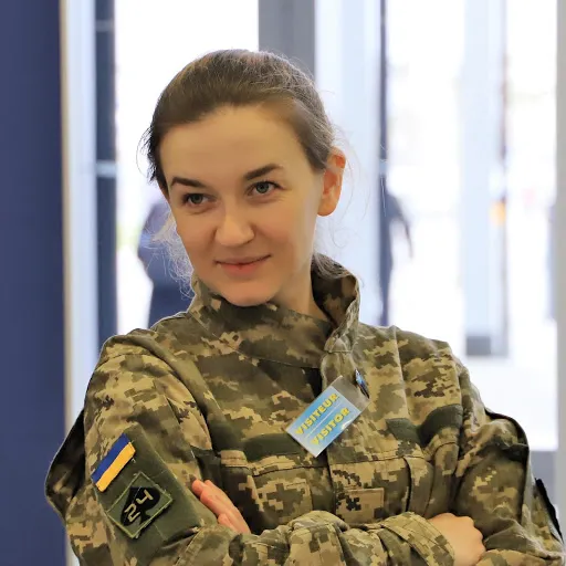 Андріана Сусак-Арехта в піксельному камуфляжі зі схрещеними руками й легкою усмішкою, на грудях бейдж «Visiteur / Visitor»