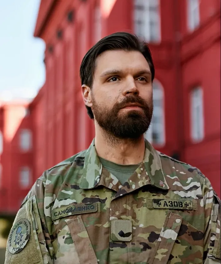 Ілля Самойленко з бородою, у камуфляжі з нашивками «Самойленко» та «Азов», на тлі червоного корпусу Київського університету