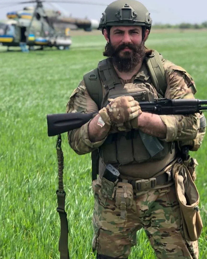 Максим Яловцов у військовій формі та шоломі зі зброєю, позаду гелікоптер