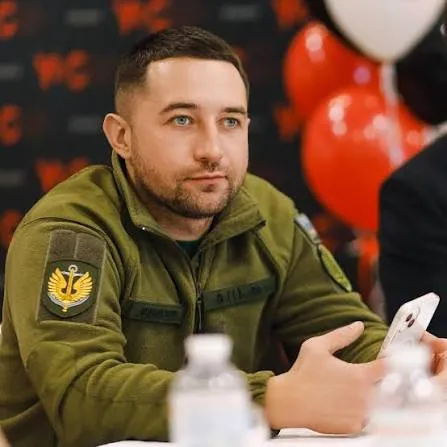 Роман Грибов у військовому фліску з шевроном на рукаві сидить за столом із телефоном у руці